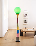 Green-globe Clérachic floor lamp with a multicolour stem in a white room with a low wooden table and pale floorboards.