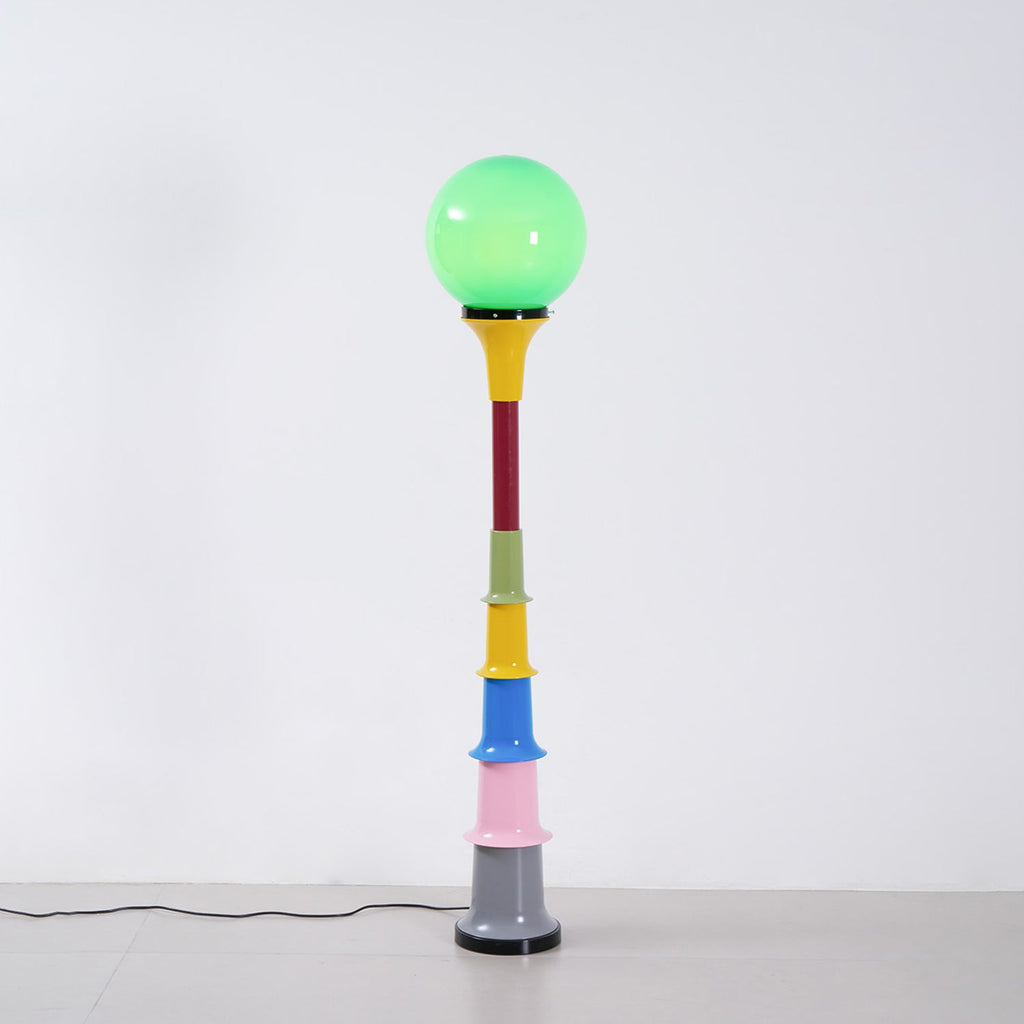 Green-globe Clérachic floor lamp with a yellow funnel and multicolour stem, standing on a pale floor by a white wall.