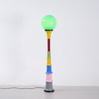 Green-globe Clérachic floor lamp with a yellow funnel and multicolour stem, standing on a pale floor by a white wall.