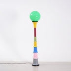 A Clérachic floor lamp with a lit green globe and stacked multicolour tiers, standing against a light grey wall.