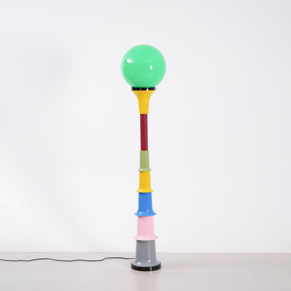 A Clérachic floor lamp with a lit green globe and stacked multicolour tiers, standing against a light grey wall.