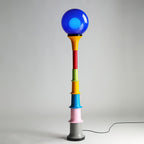 Blue-globe Clérachic floor lamp with a yellow funnel and multicolour tiers on a grey studio backdrop.