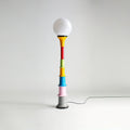White-globe Clérachic floor lamp with a multicolour stacked stem and grey foot, on a light grey backdrop.