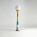 White-globe Clérachic floor lamp with a multicolour stacked stem and grey foot, on a light grey backdrop.