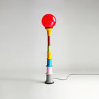 A red-globe Clérachic floor lamp with a multicolour stacked stem, on a light studio backdrop with its cord trailing.