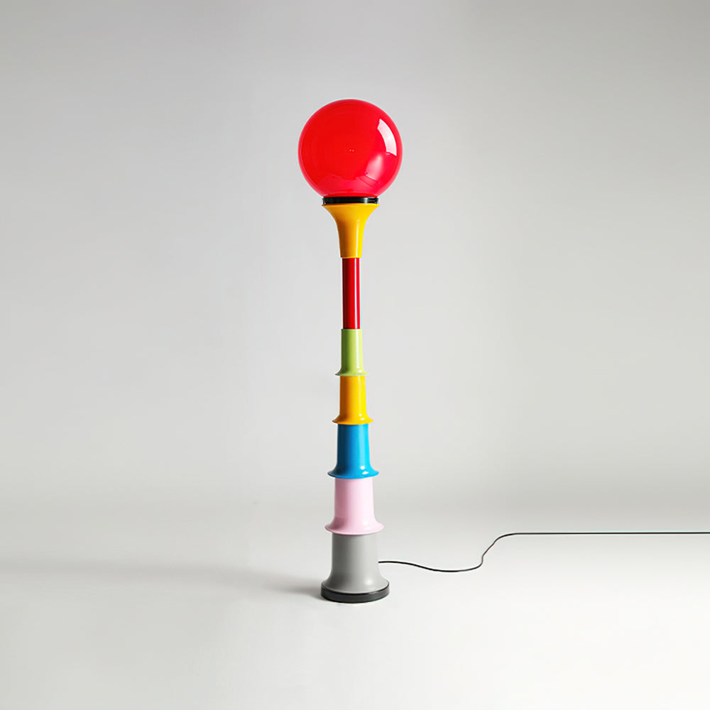 A red-globe Clérachic floor lamp with a multicolour stacked stem, on a light studio backdrop with its cord trailing.