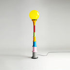 A Clérachic floor lamp with a yellow globe and a multicolour stacked stem, on a grey studio backdrop.