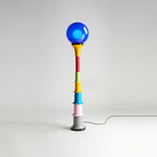 Clérachic floor lamp with a blue globe and a multicolour stacked stem, with its cord looping across the studio floor.
