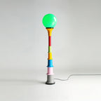 Green-globe Clérachic floor lamp with a multicolour stacked stem and a grey foot, on a studio backdrop.