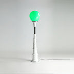 A Clérachic floor lamp with a green globe and white ribbed stem with a chrome band, on a plain studio backdrop.