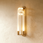 Clérachic wall light of interlocking frosted loops on a slim brass backplate, glowing warm against a limewashed wall.