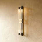 Clérachic tall wall light with two linked frosted loops and dark metal end caps, lit on a textured plaster wall.