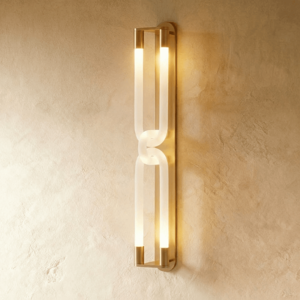 Clérachic tall wall light in brushed gold, its frosted interlocking loops lit warm along a textured plaster wall.