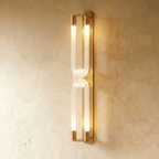 Clérachic tall wall light in brushed gold, its frosted interlocking loops lit warm along a textured plaster wall.