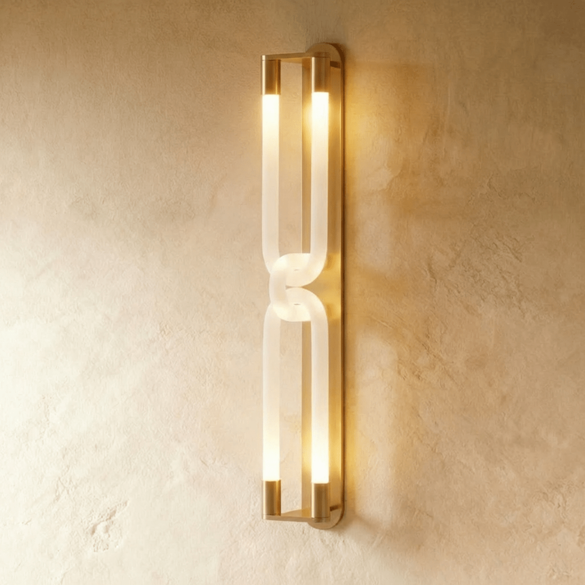 Clérachic tall wall light in brushed gold, its frosted interlocking loops lit warm along a textured plaster wall.