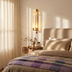 Clérachic brass wall light with frosted loops above a bedside table, in a sunlit bedroom with linen and a wool throw.