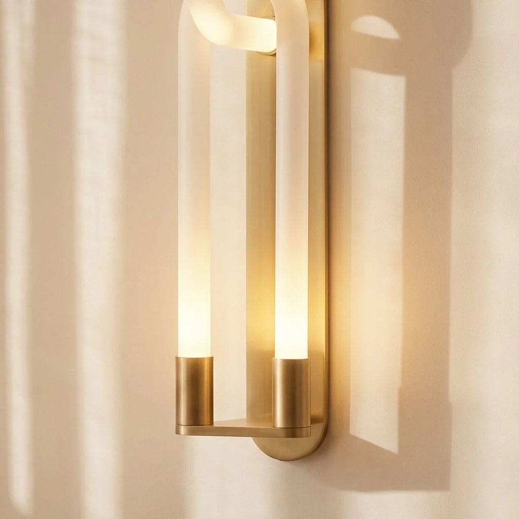 Clérachic brass wall light close up: two frosted tubes in gold cylinder bases on a slim backplate, lit warm.