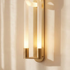 Clérachic brass wall light close up: two frosted tubes in gold cylinder bases on a slim backplate, lit warm.
