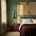 Clérachic dark bronze wall light above a burr-wood bedside table, against a green plaster wall in a bedroom.