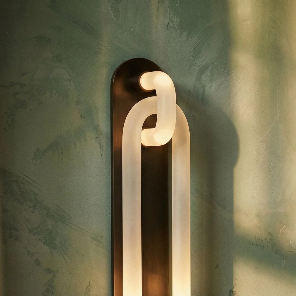 Clérachic wall light close up: frosted loops lit from within, on a dark bronze backplate against green plaster.