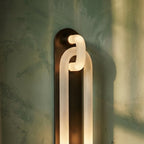Clérachic wall light close up: frosted loops lit from within, on a dark bronze backplate against green plaster.