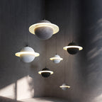 Five Clérachic planet pendants in grey and black hang at different heights against a dark textured wall, each with a glowing ring.