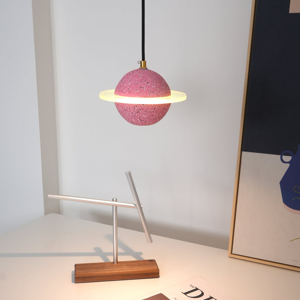 A sand pink Clérachic pendant with a glowing ring hangs above a desk with a balancing metal sculpture, a framed print and a magazine.
