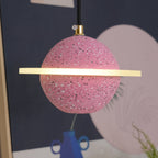 Sand pink speckled sphere with a flat glowing ring, this Clérachic pendant seen up close against a navy print with a pink circle.
