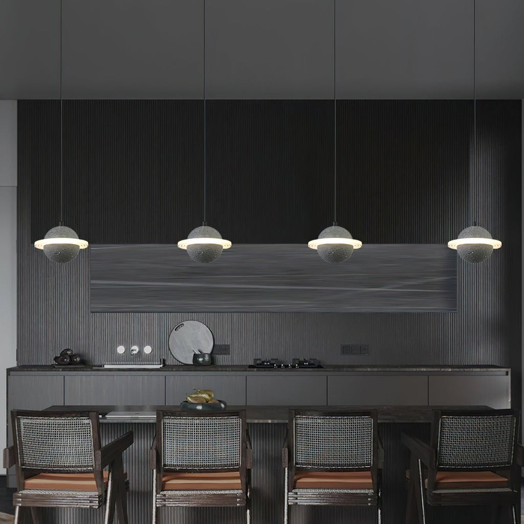 Four Clérachic planet pendants in a row above a dark kitchen island, each a pale sphere with a glowing ring, with woven chairs below.