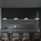 Four Clérachic planet pendants in a row above a dark kitchen island, each a pale sphere with a glowing ring, with woven chairs below.