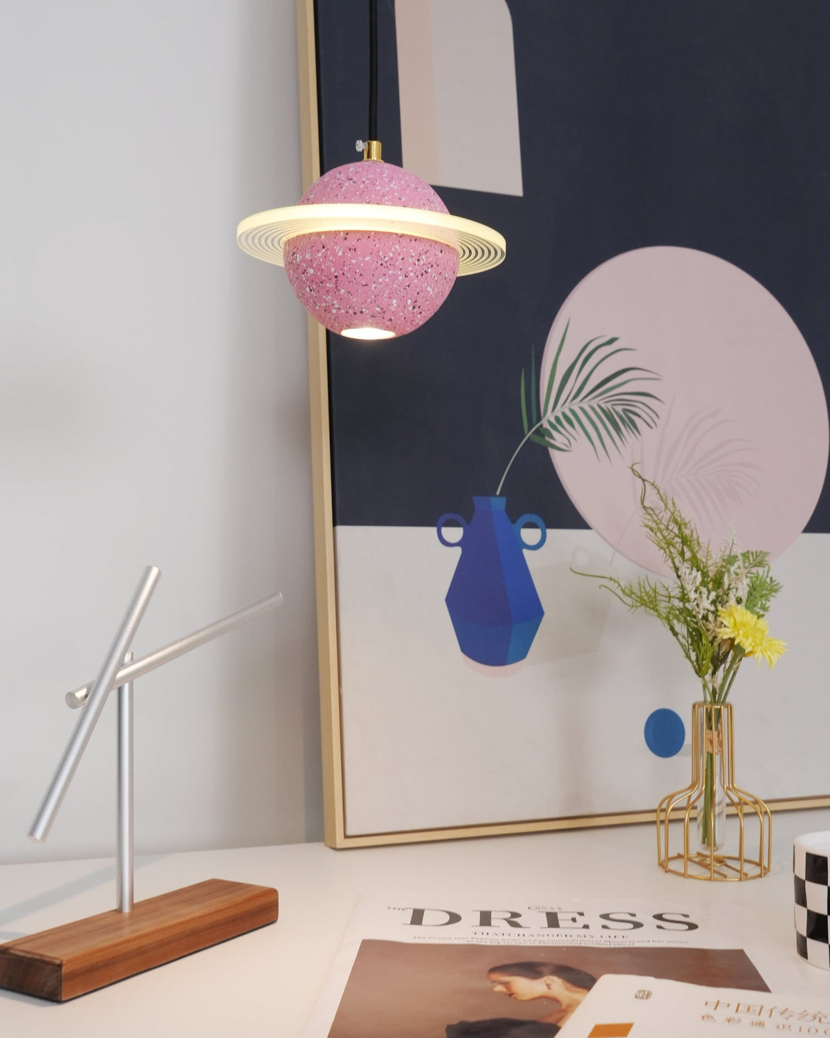 Lit sand pink Clérachic pendant with a ring, hung above a desk with a blue vase, yellow flowers, a magazine and a walnut-based sculpture.