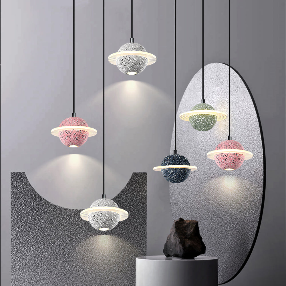 Group of Clérachic planet pendants in pink, white, green, black and grey, hanging at varied heights beside an oval mirror and a dark rock.