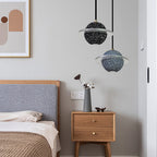 Two Clérachic planet pendants, one black speckled and one grey blue, hang beside a wood bed with a grey headboard and a bedside table.
