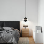 A sand black Clérachic pendant with a glowing ring hangs above a wood bedside table and a bed with grey linen, in front of a black headboard.