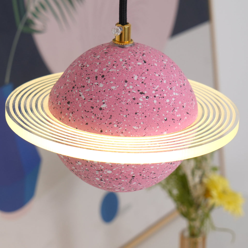 Close view of the sand pink Clérachic pendant, with a ridged glowing ring around the speckled sphere and a blue vase behind.