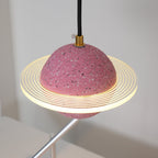Lit sand pink Clérachic pendant with a ridged ring and brass cord top, seen against a pale wall with a metal sculpture arm behind.