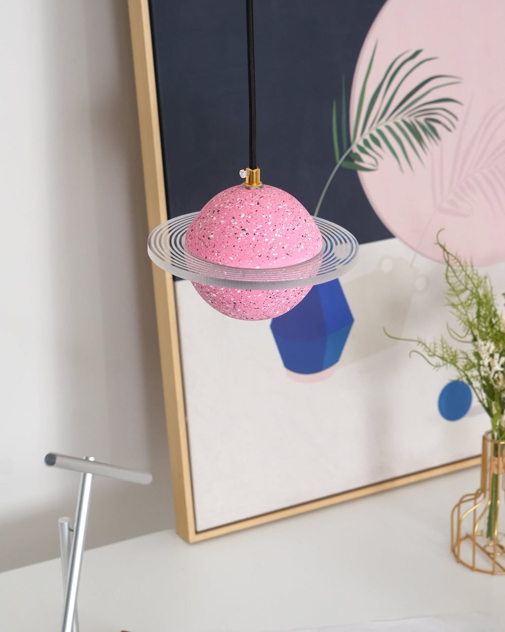 Unlit sand pink Clérachic pendant with a clear ring hangs in front of a gold-framed print with a blue vase and a vase of yellow flowers.