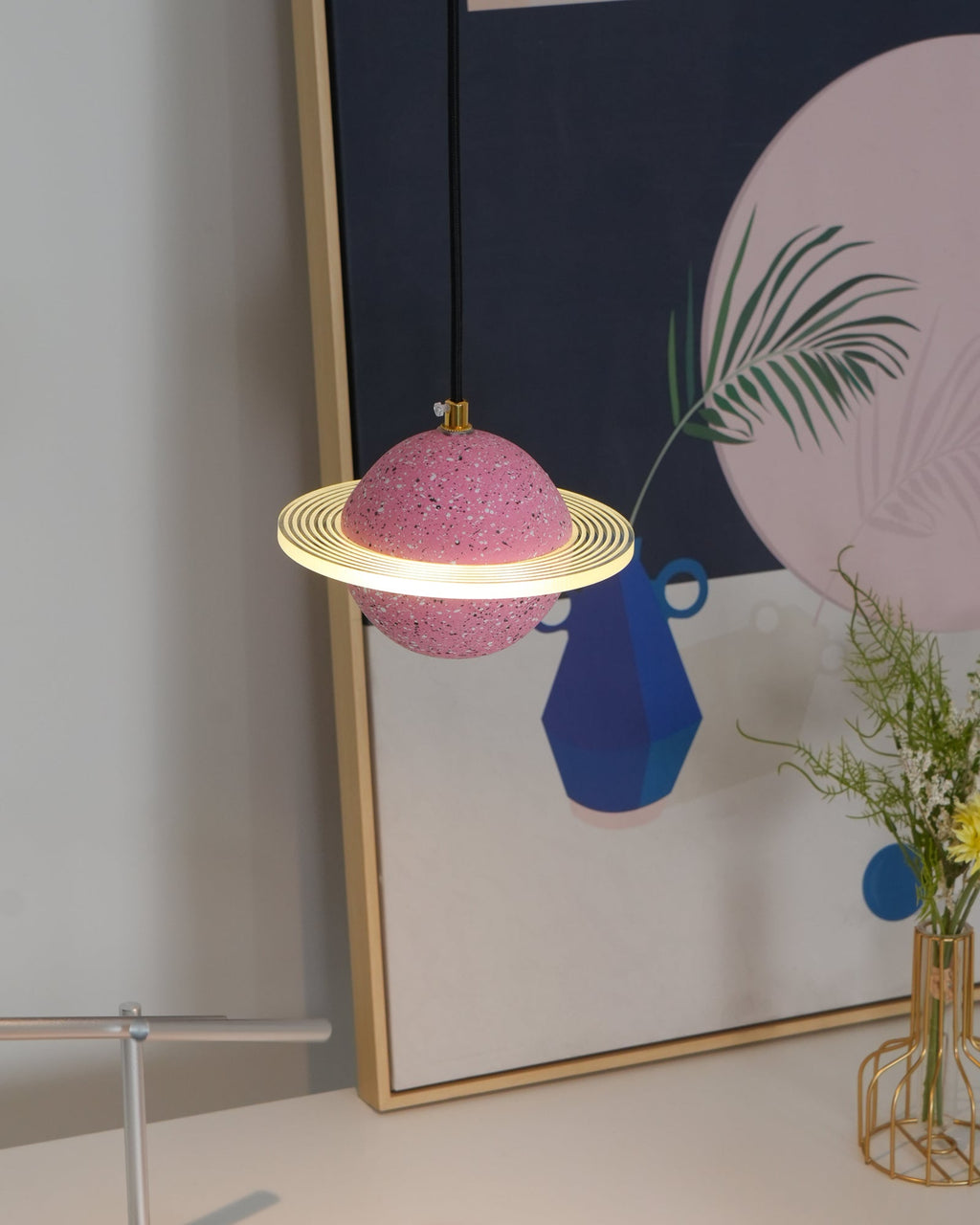 A Clérachic pendant with a sand pink sphere and a glowing flat ring hangs before a framed print with a blue vase and green fronds.