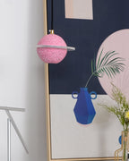 A sand pink Clérachic pendant with a flat, unlit ring hangs beside a tall framed print with a blue vase and a pink circle.