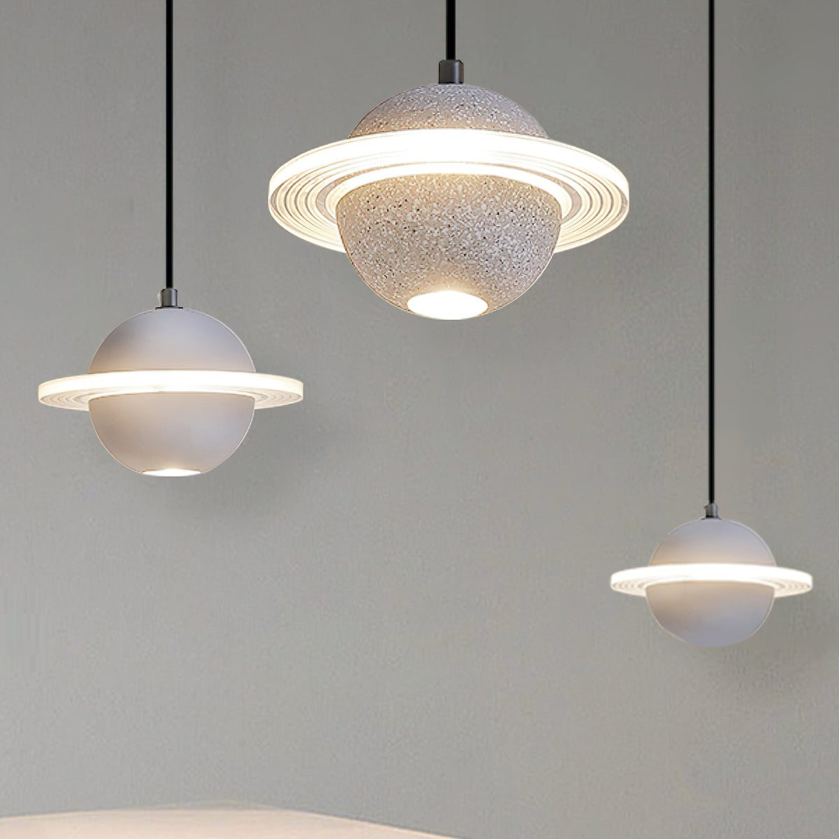 Three Clérachic planet pendants in grey and white hang at different heights against a grey wall, with glowing rings and downlights.