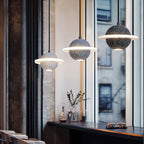 Three Clérachic planet pendants, from pale grey to black, hang beside a tall window onto a city street, above a dark counter.