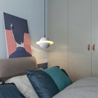 A small white Clérachic pendant with a glowing ring hangs by a bed with blue cushions, a pink and navy print and a pale grey wardrobe.