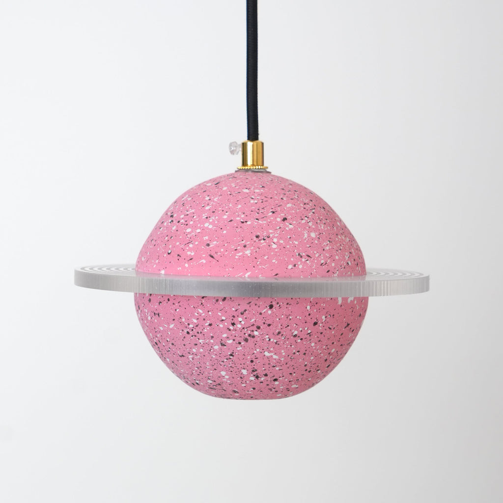 Unlit sand pink speckled sphere with a flat clear ring, hanging from a black cord with a brass top, this Clérachic pendant on pale grey.