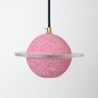 Unlit sand pink speckled sphere with a flat clear ring, hanging from a black cord with a brass top, this Clérachic pendant on pale grey.