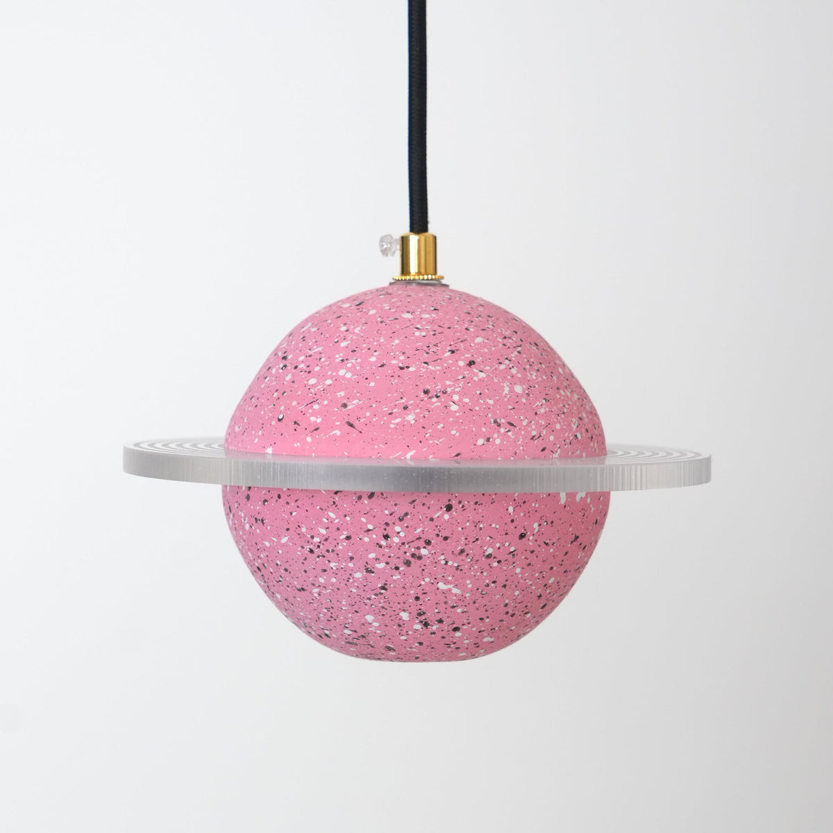 Unlit sand pink speckled sphere with a flat clear ring, hanging from a black cord with a brass top, this Clérachic pendant on pale grey.