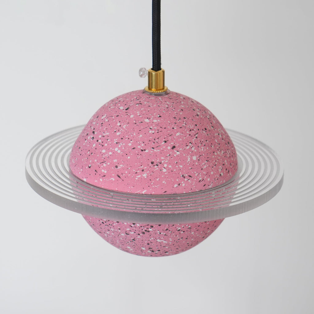 Angled view of the sand pink Clérachic pendant, with a clear ridged ring around its speckled sphere and a brass cord top.