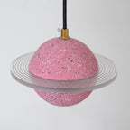 Angled view of the sand pink Clérachic pendant, with a clear ridged ring around its speckled sphere and a brass cord top.