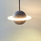 Sand black speckled Clérachic pendant with a bright glowing ring, hanging on a fine cord against a warm cream wall.