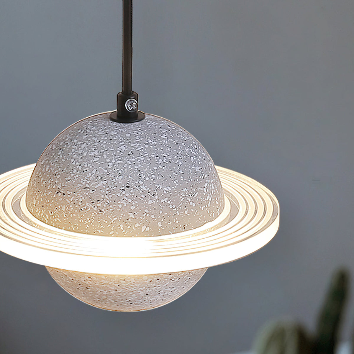 Close view of a pale speckled Clérachic pendant with its glowing ridged ring and black cord top, against a blue-grey wall.