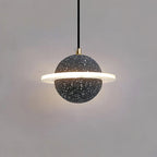 Sand black speckled Clérachic pendant with a brass cord top and a bright ring, lit against a grey wall.